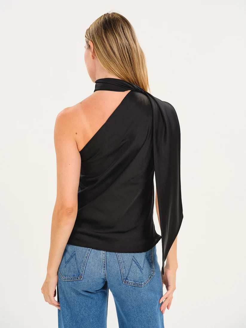 A woman shown from the back wearing a black one-shoulder draped top with a long tie detail and blue jeans, photographed against a plain white studio background.