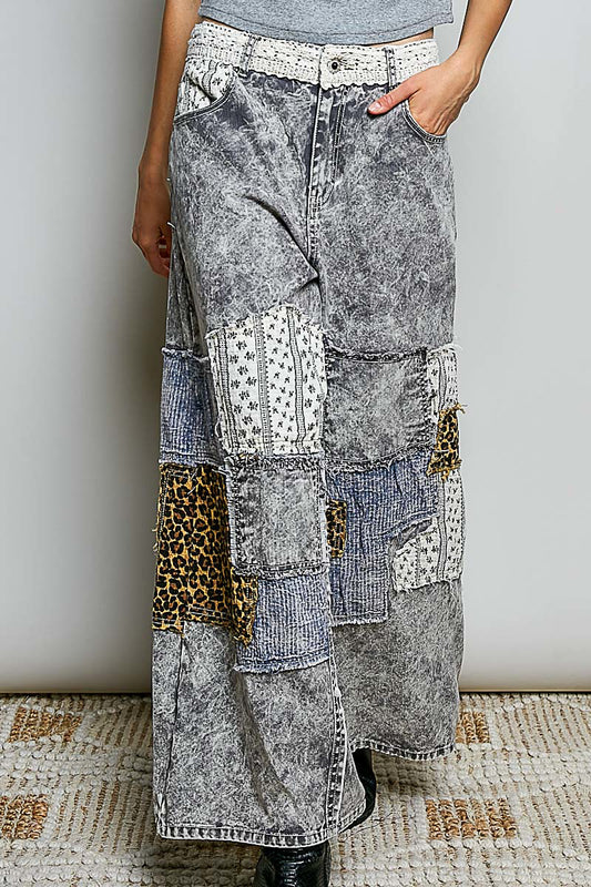 A person wearing a long patchwork denim skirt with acid-wash gray denim, patterned fabric panels, and leopard print patches, paired with a gray top and black boots.