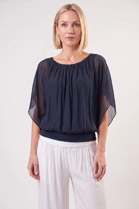 A woman modeling a sheer black short-sleeve blouse with a gathered neckline and white wide-leg pants, standing against a plain light gray studio background.