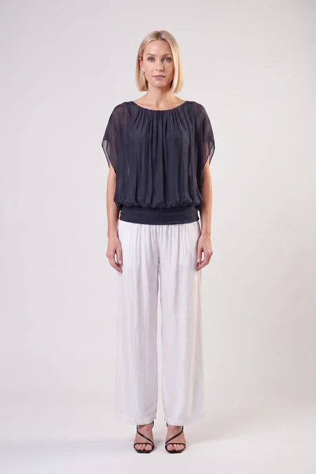 A woman modeling a sheer black short-sleeve top with a gathered neckline, paired with white wide-leg pants and black strappy heels, against a plain light gray studio background.
