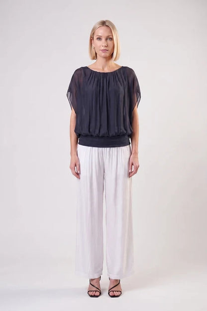 A woman modeling a sheer black short-sleeve top with a gathered neckline, paired with white wide-leg pants and black strappy heels, against a plain light gray studio background.