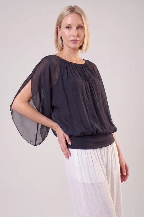 A woman modeling a dark olive green sheer draped blouse with wide flutter sleeves and a gathered neckline, paired with white pleated trousers, against a light gray studio background.