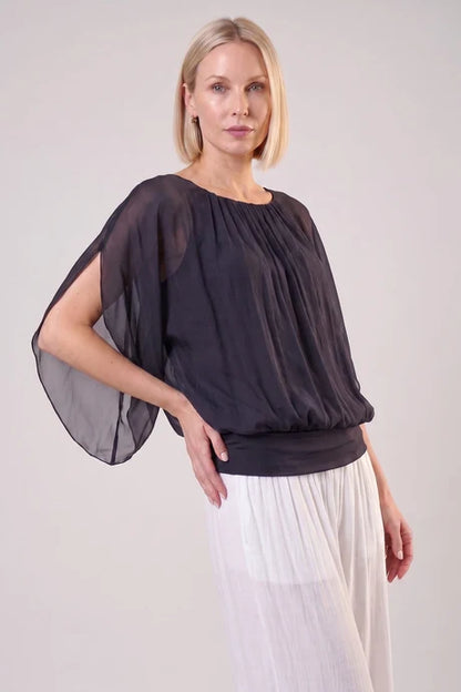 A woman modeling a dark olive green sheer draped blouse with wide flutter sleeves and a gathered neckline, paired with white pleated trousers, against a light gray studio background.