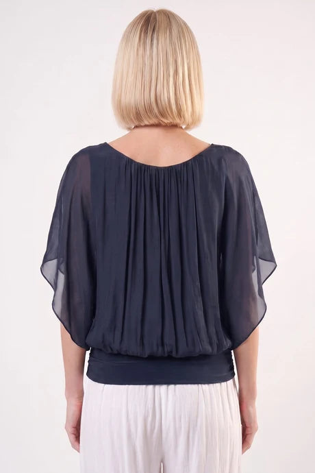A woman viewed from the back wearing a sheer dark navy blue draped blouse with wide flutter sleeves and white pants, against a plain white studio background.