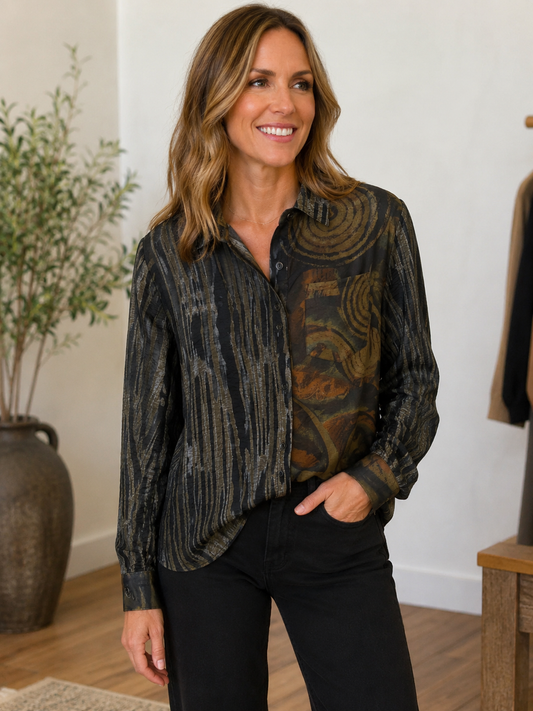 A woman wearing a long-sleeve button-up shirt with a black, brown, and gold abstract pattern, paired with black pants, standing in a brightly lit indoor setting.