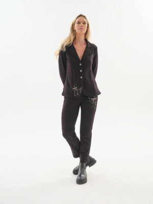 A woman modeling a black button-front jacket and matching black pants with decorative silver embellishments, standing against a plain white studio background.