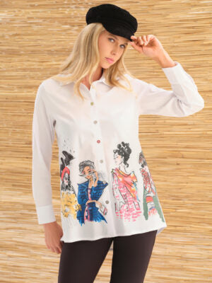 A woman wearing a white long-sleeve button-up shirt with a colorful fashion illustration print on the front, paired with black pants and a black beret, posed against a light wood slat wall.