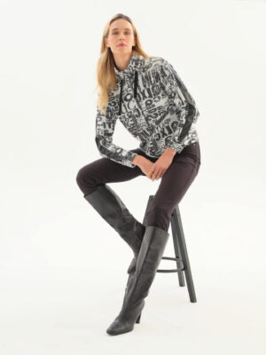 A woman sits on a black stool wearing a black and white patterned long-sleeve blouse, dark brown pants, and tall black heeled boots against a plain white studio background.